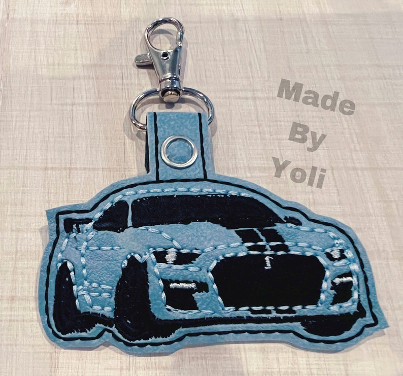K&ouml;nnte beinhalten: Ein hellblauer Schl&uuml;sselanh&auml;nger aus Filz mit einem schwarzen gestickten Bild eines Autos. Der Schl&uuml;sselanh&auml;nger hat einen silbernen Metallring und einen Clip. Der Text "Made By Yoli" ist im Hintergrund sichtbar.