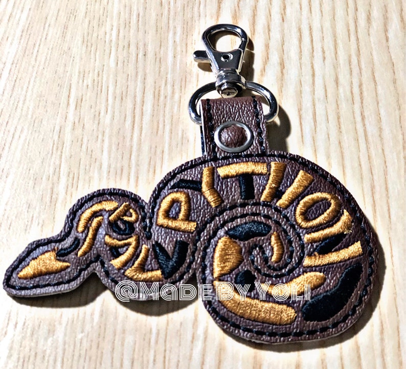 May include: Brown leather keychain featuring a gold and black embroidered snake design. The snake is curled up with the word "Revolution" embroidered around it. The keychain has a silver metal ring and a silver metal clasp.