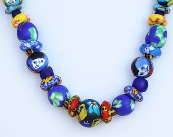 Peut inclure: Un collier coloré composé de perles de verre bleues, jaunes, rouges et blanches. Les perles sont décorées de motifs variés, notamment des visages et des fleurs.