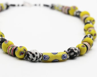 Peut inclure: Un collier composé de perles de verre colorées. Les perles sont principalement jaunes, avec des accents noirs, blancs et rouges. Le collier est enfilé sur un cordon et a un design simple.