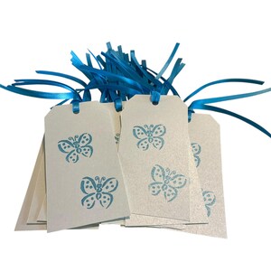 May include: A stack of off-white gift tags with a blue butterfly design. Each tag is adorned with a blue ribbon and blue fringe. The tags are perfect for adding a personalized touch to gifts or crafts.