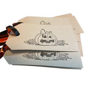 May include: A stack of light brown gift tags with black ink illustrations of a jack-o'-lantern and Snoopy. The tags have black and orange ribbon attached. The tags are for Halloween.