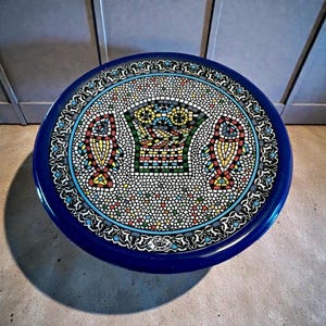 May include: A round, mosaic-tiled table with a vibrant blue border. The mosaic features a central basket design flanked by two fish motifs, all in a colorful array of tiles. The table's diameter is approximately 24 inches.