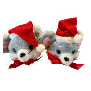 May include: Two plush mouse slippers featuring soft gray fur, white muzzles, and large pink-lined ears. Each mouse wears a bright red felt Santa hat with a white pom-pom and a matching red satin ribbon bow tied around its neck.