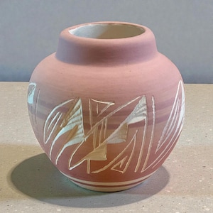 May include: A small, round, pink ceramic vase with a wide opening. The vase features a carved design of abstract shapes and lines in white. The base has two white stripes. The interior is white.