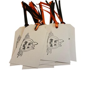 May include: A stack of cream-colored gift tags with a black and white pumpkin design. Each tag has a black and orange ribbon attached for hanging. The tags are perfect for Halloween gifts.