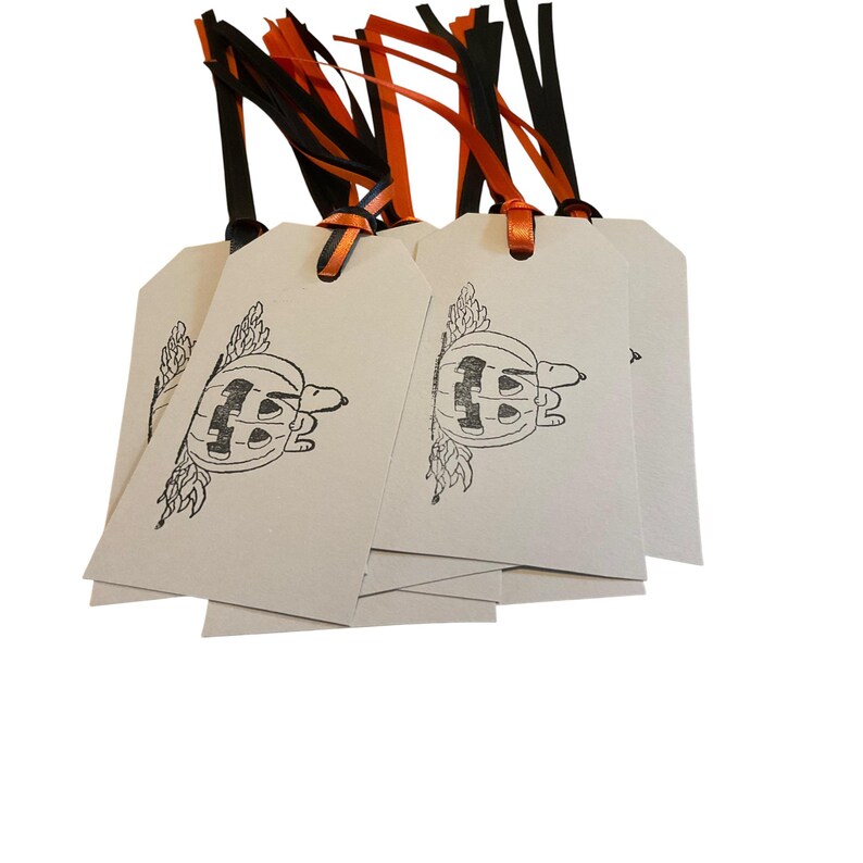 May include: A stack of cream-colored gift tags with a black and white pumpkin design. Each tag has a black and orange ribbon attached for hanging. The tags are perfect for Halloween gifts.