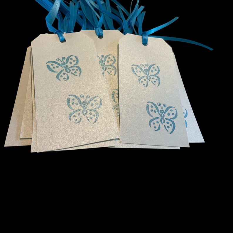 May include: A stack of off-white gift tags with a blue butterfly design. Each tag is strung with a blue ribbon. The tags are arranged in a fan shape against a black background. The butterfly design is repeated on each tag.