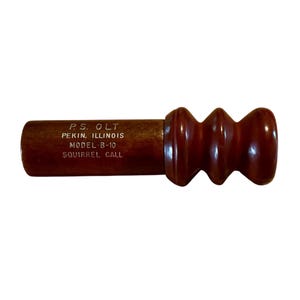 May include: A brown wooden squirrel call with a bulbous end and the text "P.S. OLT PEKIN, ILLINOIS MODEL-B-10 SQUIRREL CALL" printed on the side. The call has a smooth, polished finish and is designed for hunting.