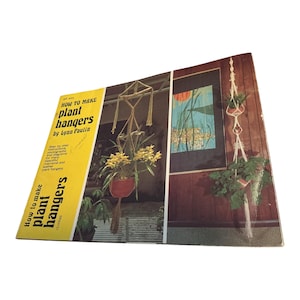 May include: A softcover instructional book titled How to Make Plant Hangers by Lynn Paulin. The cover features a bright yellow side panel with black text and a photographic display of various macrame plant hangers holding potted green plants.