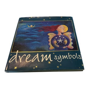 May include: A hardcover book titled dream symbols with a dark blue cover featuring a celestial illustration of a figure with long red hair, wings, and a moon face, surrounded by stars and a crescent moon against a deep blue night sky background.