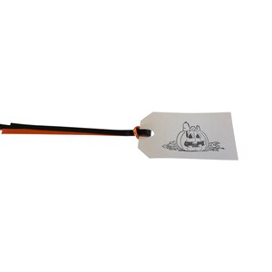 May include: A white Halloween gift tag with a black and white illustration of a jack-o'-lantern and a character. The tag is attached to a black and orange ribbon. The tag is rectangular with a rounded top.