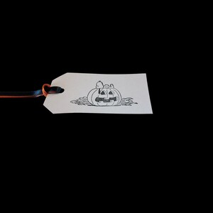 May include: A white tag with a black and orange ribbon. The tag features a black illustration of a jack-o'-lantern with a cartoon character on top. The jack-o'-lantern is surrounded by leaves.