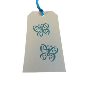 May include: A rectangular gift tag with two teal butterfly designs. The tag is a light tan color and has a teal ribbon attached at the top. The butterflies are stylized with dotted details.