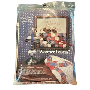 May include: A patchwork kit by Yours Truly, featuring a framed image with the text "Quilters make Warmer Lovers." The kit includes fabric pieces for a 13-inch square quilt, with a decorative lamp in the foreground. The packaging is sealed in plastic.