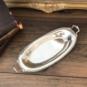May include: An oval silver-toned serving tray with a polished, reflective surface and a decorative, textured border. The tray features two ornate, scrolled handles on opposite ends, providing a classic look for serving small items or displaying objects.