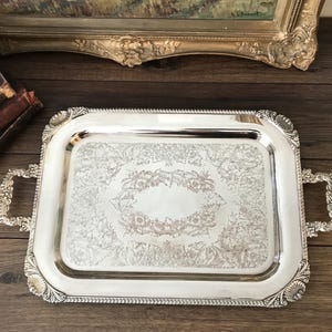 Peut inclure: Un plateau de service rectangulaire de couleur argentée présentant des motifs floraux en relief ornés au centre et sur les bords. Le plateau comprend deux poignées décoratives sur les côtés et un détail de bordure en corde sur tout le périmètre.