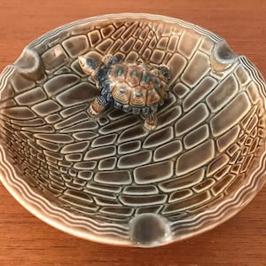 May include: A brown ceramic ashtray with a raised, textured design resembling crocodile skin. A small, brown ceramic turtle figurine sits in the centre of the ashtray.