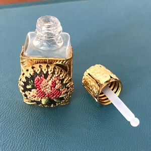 Op de afbeelding: Een klein, doorzichtig glazen flesje met een gouden metalen dop en een bloemenmotief. Het flesje heeft een druppelaar aan de dop.