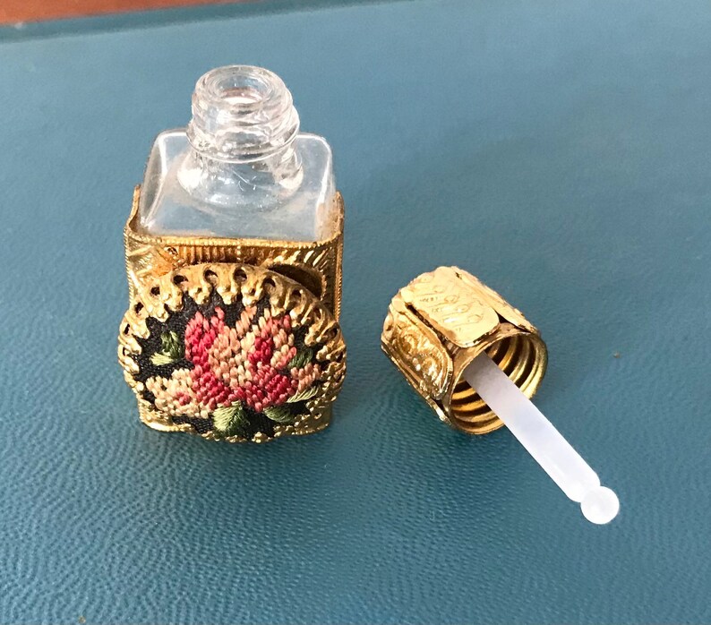 Op de afbeelding: Een klein, doorzichtig glazen flesje met een gouden metalen dop en een bloemenmotief. Het flesje heeft een druppelaar aan de dop.