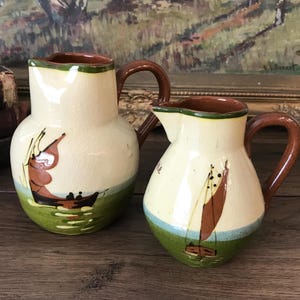 May include: Two cream-colored ceramic pitchers with brown handles and rims. Each pitcher features a hand-painted scene of a sailboat on green and blue water. The larger pitcher is on the left, and the smaller pitcher is on the right, both displaying a glossy finish and a rounded body shape with a distinct pouring spout.