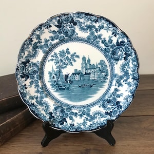 May include: A blue and white decorative plate featuring a floral pattern and a central landscape scene. The plate has a scalloped edge and a gold trim. The landscape depicts a town with buildings, trees, and boats. The plate is displayed on a black stand.