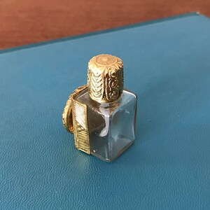 Op de afbeelding: Een klein, helder glazen flesje met een gouden dop. De dop heeft een ingewikkeld ontwerp en het flesje heeft een vierkante vorm.