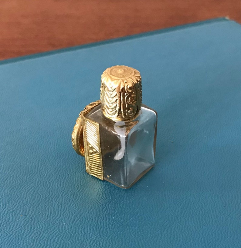 Op de afbeelding: Een klein, helder glazen flesje met een gouden dop. De dop heeft een ingewikkeld ontwerp en het flesje heeft een vierkante vorm.