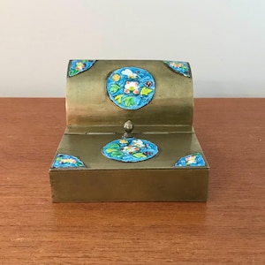 May include: A brass inkwell with a hinged lid and blue enamel floral designs. The inkwell has a small knob on the top.