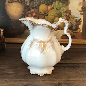 May include: White ceramic pitcher with a scalloped rim and handle, accented with gold trim. The pitcher has a bulbous body and a decorative, ornate design. The background includes a framed painting of fruit.