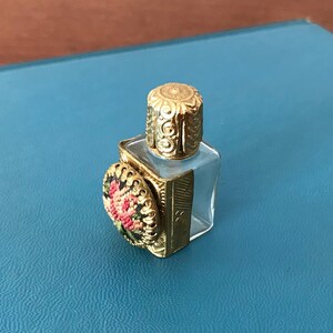 Op de afbeelding: Een klein, helder glazen flesje met een gouden dop en een bloemenmotief aan de zijkant. Het flesje is versierd met een gouden metalen frame.