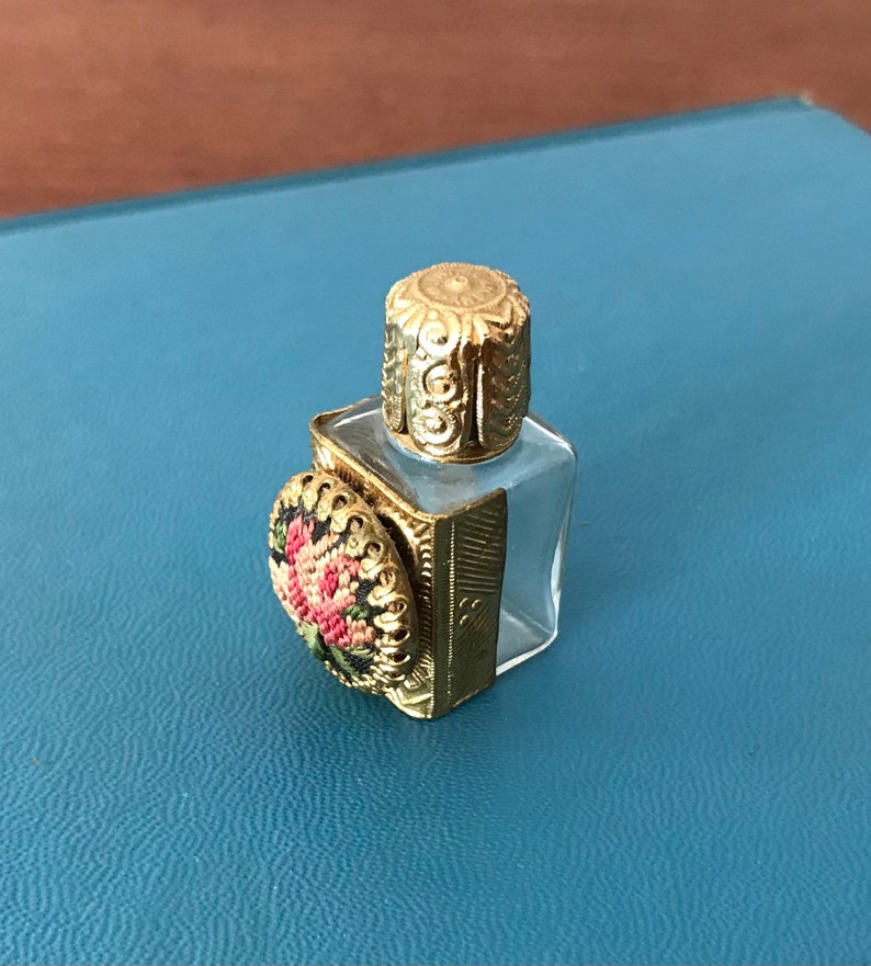 Op de afbeelding: Een klein, helder glazen flesje met een gouden dop en een bloemenmotief aan de zijkant. Het flesje is versierd met een gouden metalen frame.