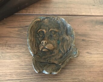 May include: A bronze-coloured, dog-shaped decorative item. The dog's face is detailed with visible eyes, nose, and fur. The item has a collar and is likely made of metal. The overall appearance suggests an antique or vintage piece.
