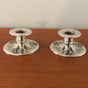May include: Two silver candle holders with a decorative, openwork design. The holders are on a wooden surface.