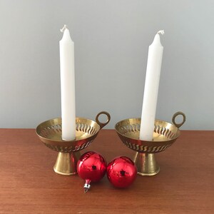 Peut inclure: Deux bougeoirs en laiton avec des bougies blanches et deux boules de Noël rouges sur une surface en bois.