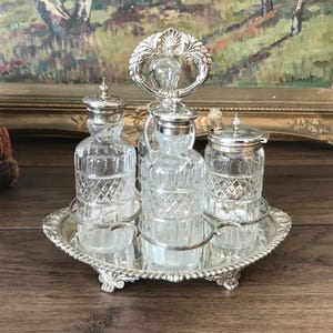 May include: A silver-plated condiment set featuring a mirrored oval tray with ornate edges and a central handle. It holds three clear glass bottles with diamond-cut patterns and silver-plated lids, including one with a small silver spoon.