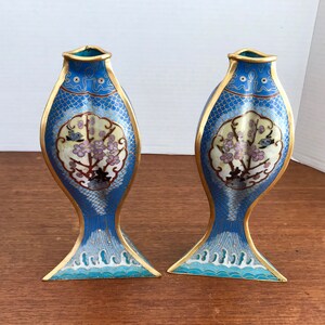 May include: A pair of blue and gold cloisonn&eacute; vases shaped like fish with floral designs. The vases are decorated with a white and gold background and have a blue and white wave pattern at the bottom.
