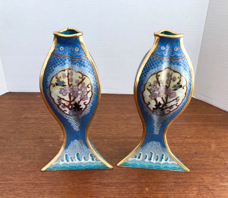 May include: A pair of blue and gold cloisonn&eacute; vases shaped like fish with floral designs. The vases are decorated with a white and gold background and have a blue and white wave pattern at the bottom.