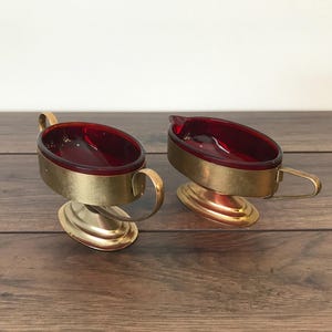 May include: A set of two vintage red glass and gold metal condiment dishes. The dishes are oval shaped and have a gold metal rim. The dishes are on a wooden surface.