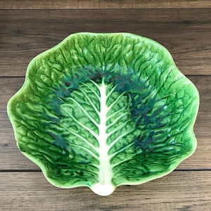 May include: A ceramic bowl shaped like a large green cabbage leaf. The bowl features a glossy finish with vibrant shades of light and dark green, highlighting the textured, ruffled edges and a prominent, smooth white central vein running through.