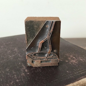 May include: Vintage letterpress printing block with a geometric design. The block is made of wood and features a copper-colored metal design. The design includes a triangular shape and a rectangular base with intricate details. The block is likely used for printing.