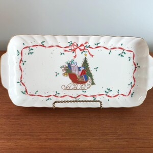 May include: White rectangular serving tray with a gold rim and a Christmas scene featuring a red sleigh with presents, a green Christmas tree, and red and green ribbon accents.