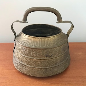 Op de afbeelding: Een vintage koperen pot met een sierlijk ontwerp en een handvat. De pot is versierd met ingewikkelde bloem- en geometrische patronen.