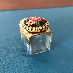 Op de afbeelding: Een klein, helder glazen potje met een gouden deksel. Het deksel is versierd met een bloemmotief dat is geborduurd in roze, wit en groen garen.