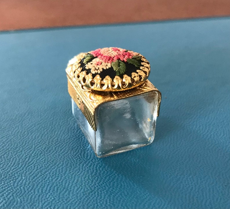 Op de afbeelding: Een klein, helder glazen potje met een gouden deksel. Het deksel is versierd met een bloemmotief dat is geborduurd in roze, wit en groen garen.