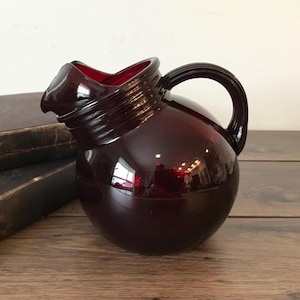 May include: A deep red glass jug with a rounded form and a handle. The jug has a spout and a ribbed neck. The jug is on a wooden surface next to a stack of books. The jug is a decorative item.