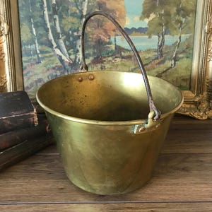 May include: A polished brass bucket with a tapered cylindrical body and a wide open top. The bucket features a dark, weathered metal swing handle attached to the sides with riveted brass brackets. The surface shows subtle signs of age and patina.