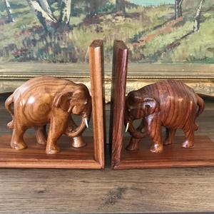 May include: Wooden elephant bookends. Each bookend has a carved elephant with tusks, standing on a rectangular base. The wood has a warm, reddish-brown colour. The bookends are in front of a framed painting.