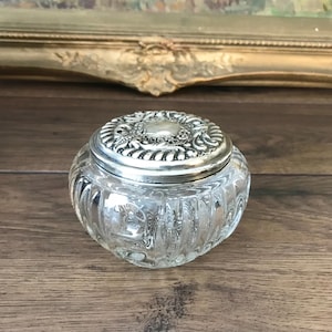 Op de afbeelding: Een doorzichtige glazen pot met een afgerond, geribbeld lichaam en een rond deksel van zilverkleurig metaal. Het deksel is voorzien van een ingewikkeld reliëfpatroon met een gladde, gepolijste schijf in het midden. De pot staat op een plat oppervlak en toont de transparante glazen constructie en de gedetailleerde metalen bovenkant.