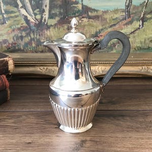 May include: A silver coffee pot with a dark gray handle and a decorative finial on the lid. The pot has a ribbed design on the lower portion and is set against a backdrop of a framed painting.